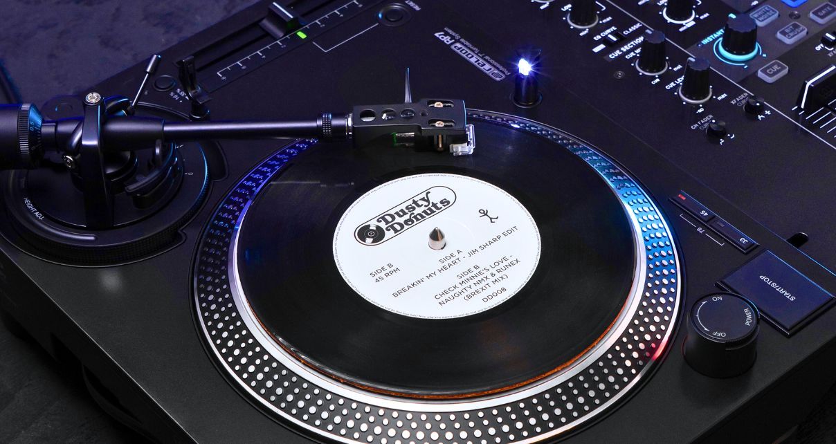 Close-up of a black Reloop RP-7 turntable. There is a black 7" single on the platter with a white label that says "Dusty Donuts". Further down the text says "Side A" and "breakin' My Heart - Jim Sharp Edit". At the bottom it says "Side B" and "Check Minnie's Love - Naughty NMX & Runex (Brexit Mix)". The tone arm is placed on the record, and the target light is shining on the record.