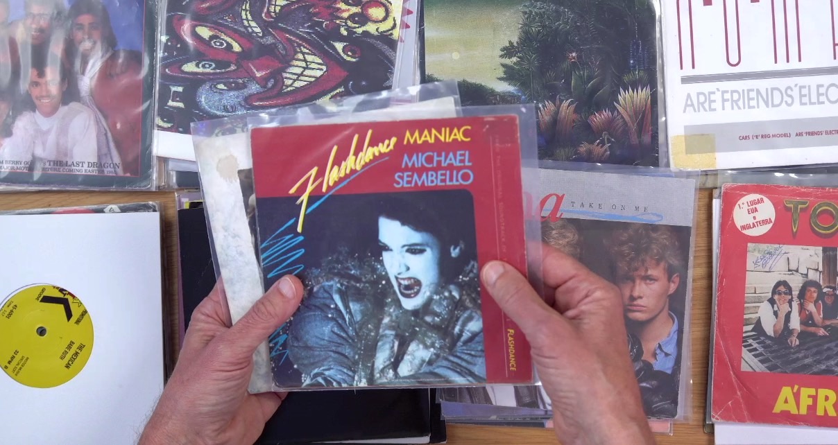 Eight piles of 7" singles sit next to each other on a wooden desk. A person holds up a stack showcasing Michael Sembello's "Maniac" single from the movie Flashdance.