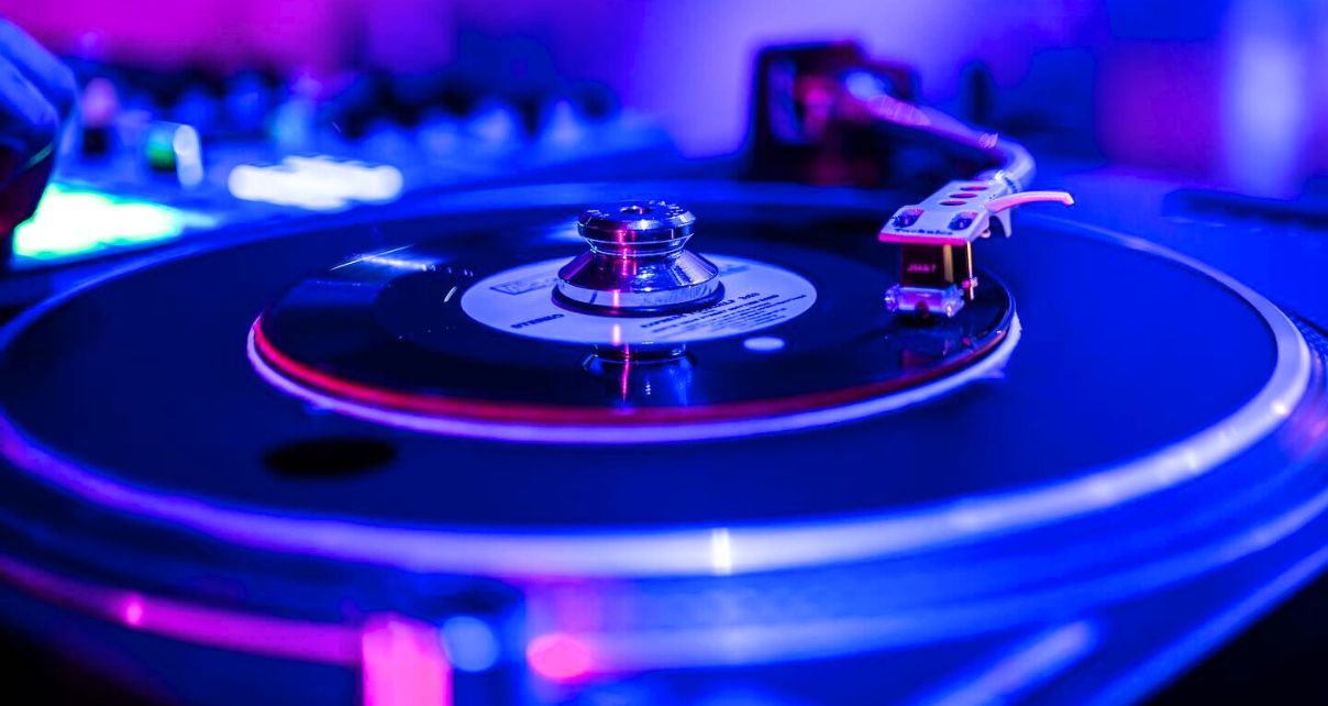 Close-up view of a 7" record on a large turntable. There is a heavyweight metal cone-shaped adapter in the centre. The whole photo is lit in blue, pink, and purple tones.