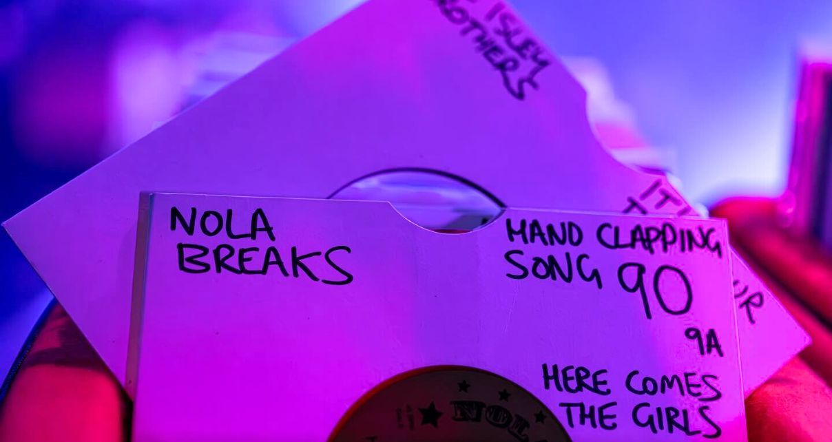 Two 7" records sit in a red bin. The records have white sleeves with writing on them. The one closest says "Nola Breaks" followed by "Hand Clapping Song" and the number 90.