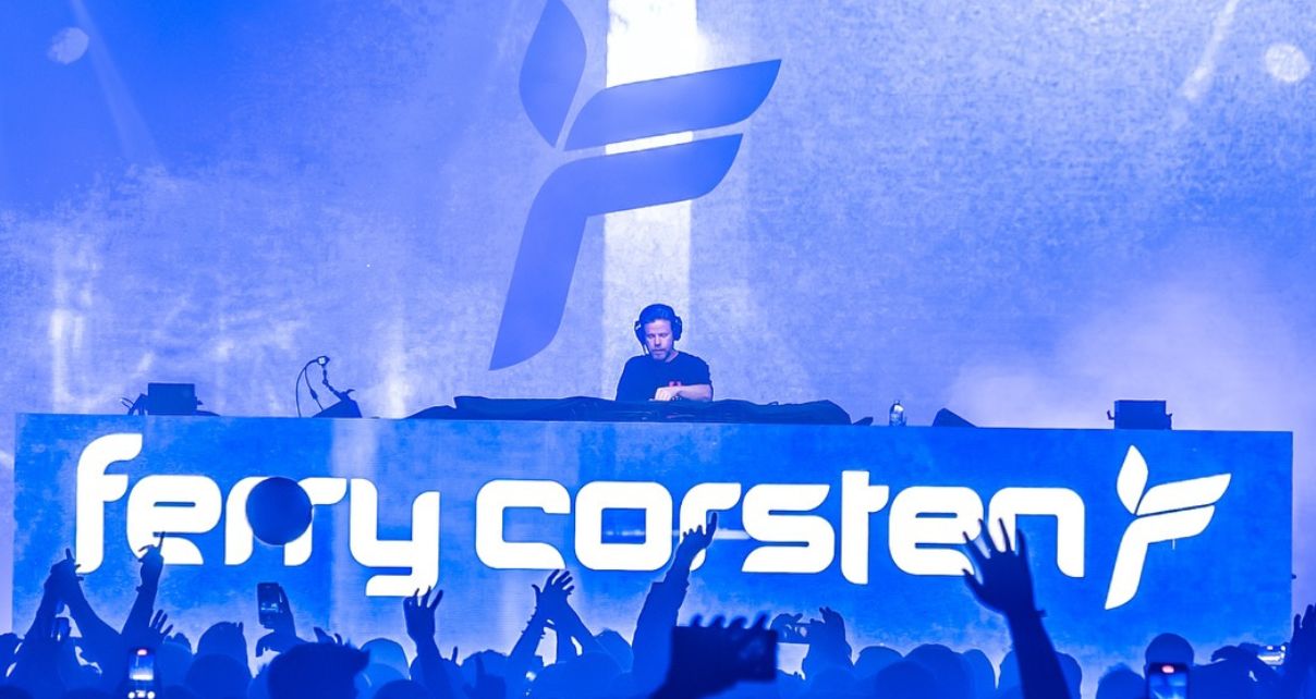 A DJ performs at a vibrant concert, with energetic fans raising their hands in front of a large illuminated sign displaying "ferry corsten."