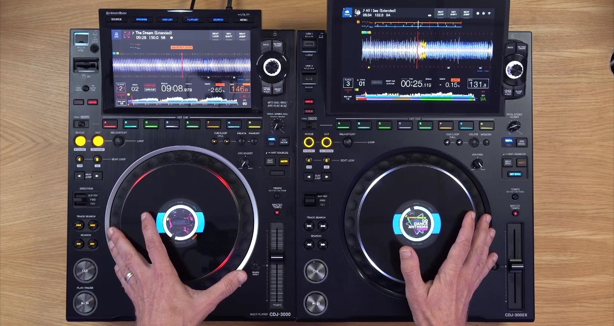 Top-down view of a CDJ-3000 media player next to a CDJ-3000X on a wooden table. A person gestures towards the players with both hands. 