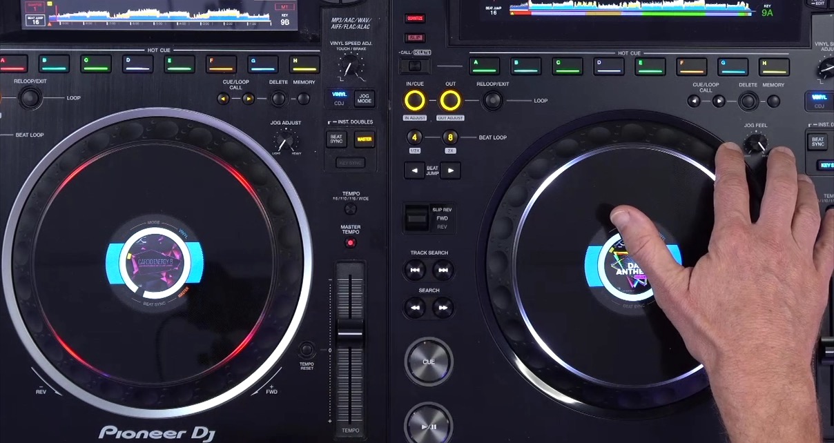 Close up of two CDJs next to each other. The one on the left is a CDJ-3000 and the one on the right is a CDJ-3000X. A person gestures towards the jog adjust knob on the right media player.