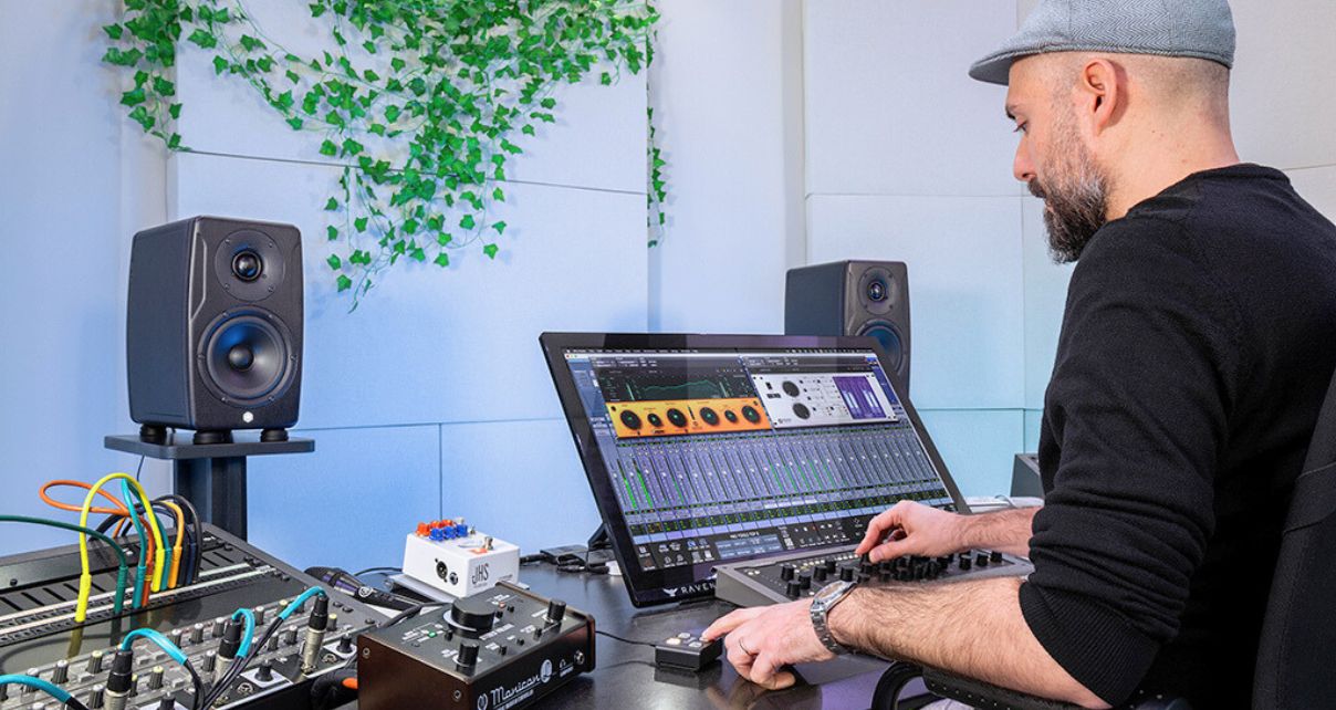 A musician uses a digital audio workstation on a large monitor, surrounded by audio equipment and studio monitors, in a well-lit space. In the background are two black IK Multimedia iLoud Precision 5 MKII studio monitors on stands, set against a white wall with green plants.