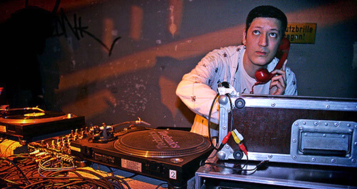 A photo of Mark Moore in a club DJing on a turntable set-up. He's looking out towards the crowd and holding a red telephone-style headphone up to his ear.