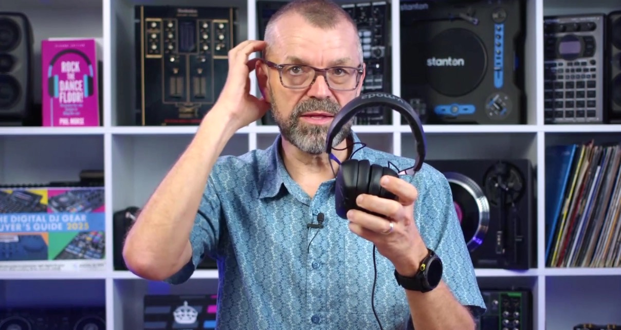 A man with close-shaven hair, glasses, and a salt and pepper beard stands in front of a bookshelf, holding a pair of black headphones up to the camera. He cups his ear with his right hand. The bookshelf behind him contains various books, DJ gear, and vinyl.