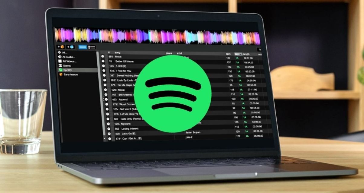 A silver laptop displaying DJ software on a wooden table. The lighting is soft and cozy, and there's a water glass set on the left side of the computer. There is a big green and black Spotify logo across the middle of the screen.