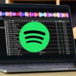 A silver laptop displaying DJ software on a wooden table. The lighting is soft and cozy, and there's a water glass set on the left side of the computer. There is a big green and black Spotify logo across the middle of the screen.
