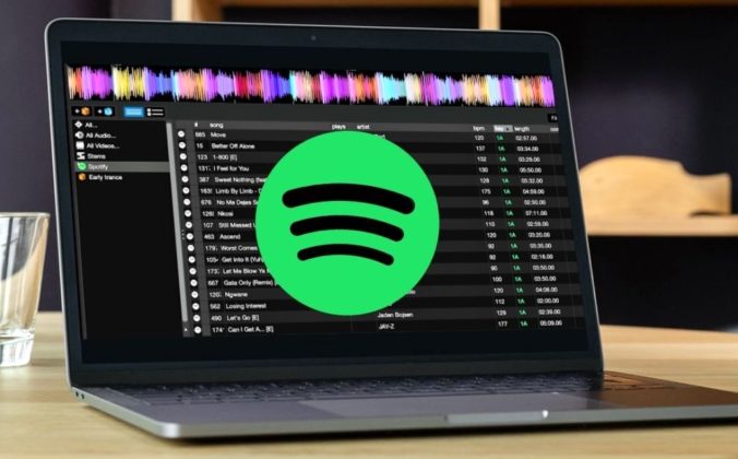 A silver laptop displaying DJ software on a wooden table. The lighting is soft and cozy, and there's a water glass set on the left side of the computer. There is a big green and black Spotify logo across the middle of the screen.