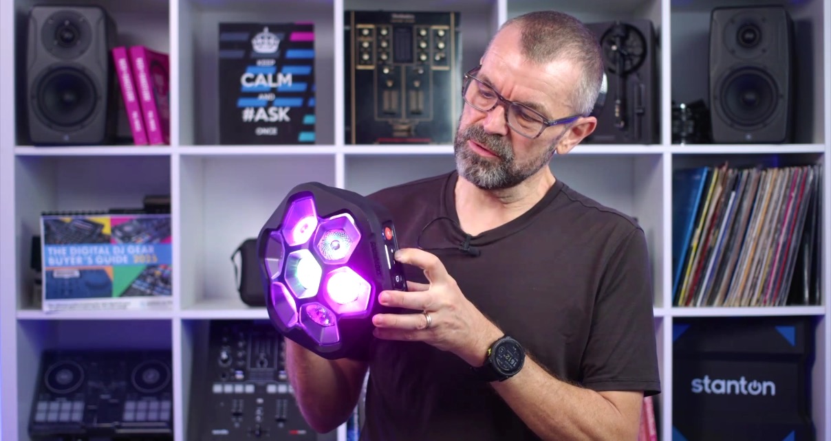 A man holds a portable Soundboks Lightboks party light in his hands and gestures towards the controls. The light is shining with pink and purple hues. The man is standing in a well-lit room with a full bookshelf behind him. 