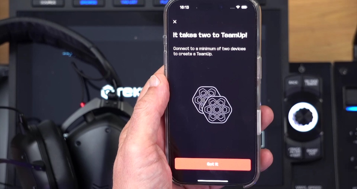 A person holds their phone above some DJ gear, showing what's displayed on the screen. The text reads "It takes two to TeamUp!" and has a small graphic showing two party lights. 