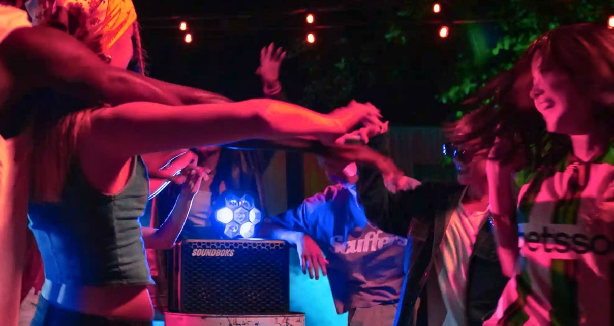 Various people dance and laugh in a vibrant outdoor setting. There's a Soundboks Go speaker and Lightboks party light in the middle. 