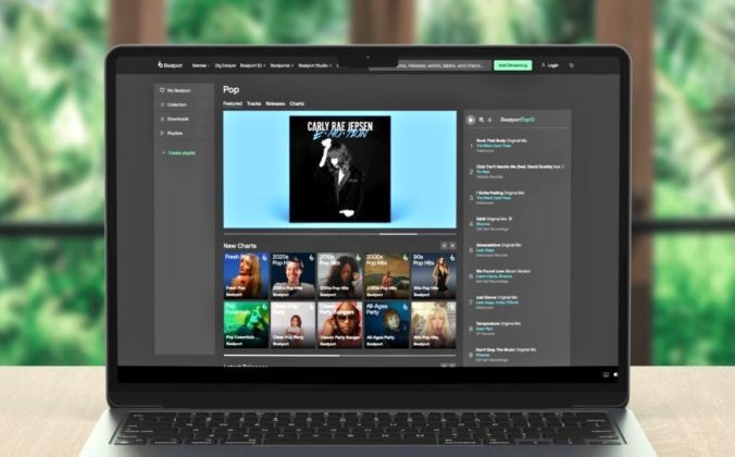 A laptop displays Beatport's pop music section, featuring Carly Rae Jepsen's album and various music charts. It sits on a light wooden desk with a blurry nature background.
