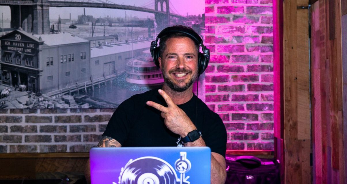 A DJ smiles at the camera and holds up a peace sign. The booth is in front of a brick wall illuminated with pink lighting, and off to the left is a black and white photograph of an old cityscape.