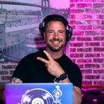 A DJ smiles at the camera and holds up a peace sign. The booth is in front of a brick wall illuminated with pink lighting, and off to the left is a black and white photograph of an old cityscape.