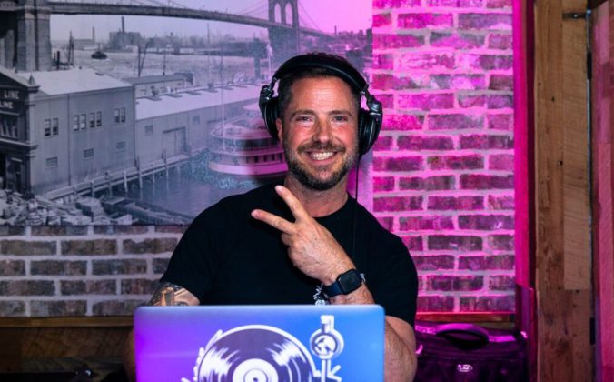A DJ smiles at the camera and holds up a peace sign. The booth is in front of a brick wall illuminated with pink lighting, and off to the left is a black and white photograph of an old cityscape.