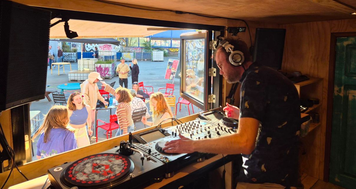 A DJ mixes on CDJs inside a wooden booth, while a lively crowd enjoys the outdoor event amidst colorful chairs and graffiti. 