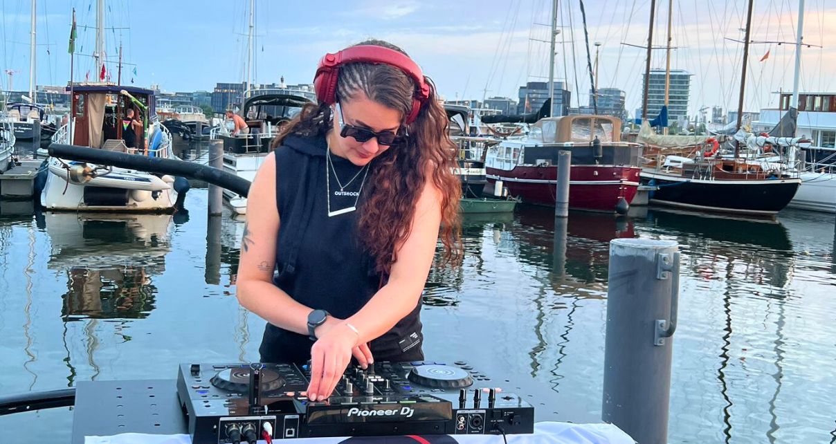 A DJ performs live at a waterfront, surrounded by moored boats and city skyline, with vibrant reflections on the water. 