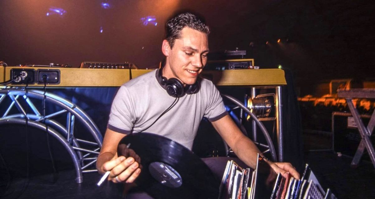 A late 90s Tiësto, smiling and facing the camera, knelt down behind the DJ booth. He's holding a cigarette and vinyl record while sorting through his collection.