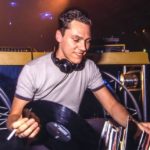 A late 90s Tiësto, smiling and facing the camera, knelt down behind the DJ booth. He's holding a cigarette and vinyl record while sorting through his collection.