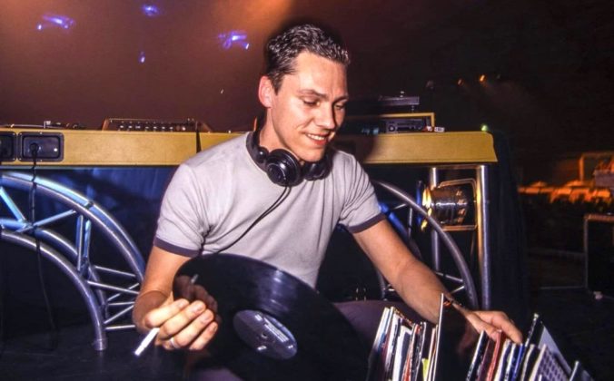 A late 90s Tiësto, smiling and facing the camera, knelt down behind the DJ booth. He's holding a cigarette and vinyl record while sorting through his collection.