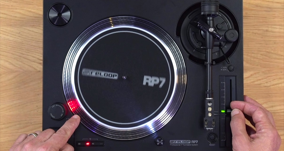 Close-up of a single black Reloop RP-7 DJ turntable on a wooden desk. A person is adjusting the pitch fader and pointing at the strobe dots.