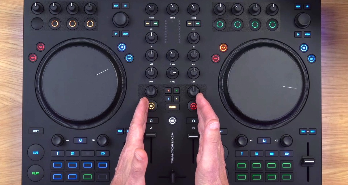 A Traktor MX2 DJ controller sits on a wooden desk in a well-lit room. A person's hands are gesturing towards the mixer section. 