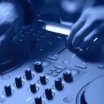 Hands adjusting the jogwheel and crossfader on a Traktor MX2 DJ controller. The whole photo has a blue tone.