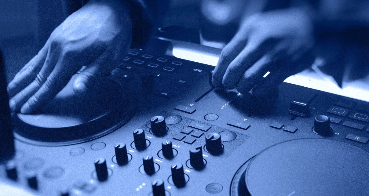 Hands adjusting the jogwheel and crossfader on a Traktor MX2 DJ controller. The whole photo has a blue tone. 