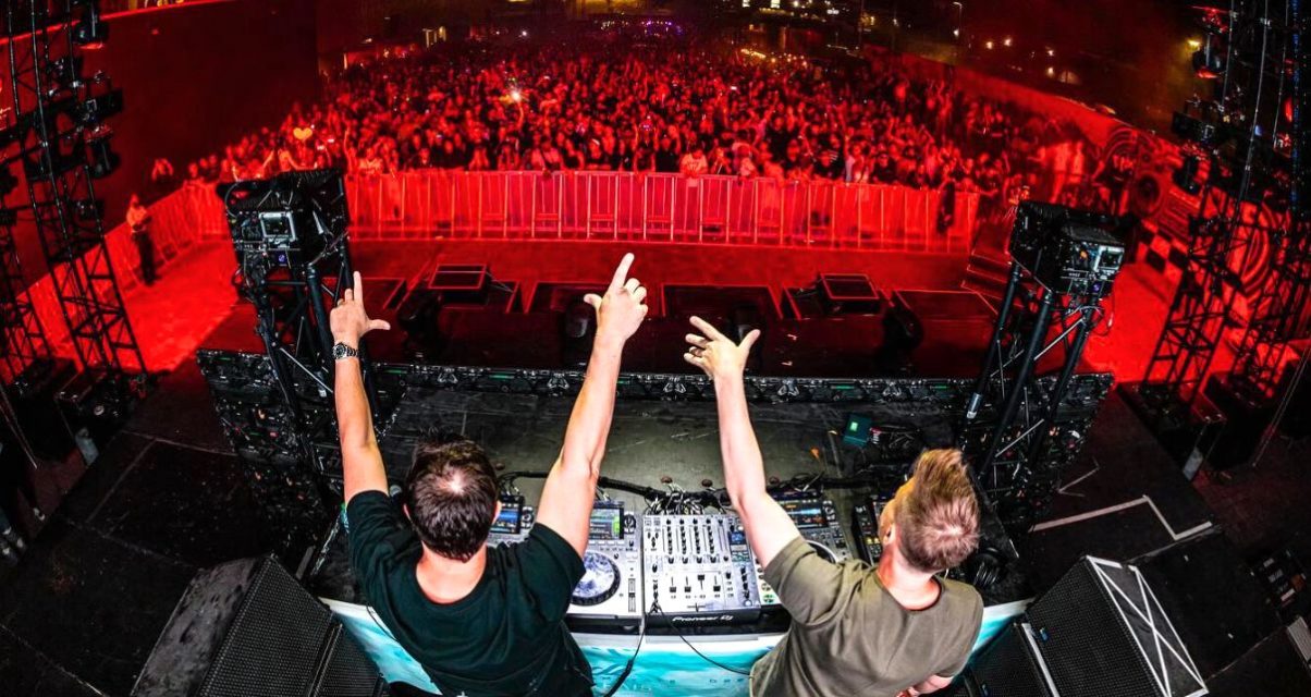 Two DJs perform on stage, raising their arms to a cheering crowd, illuminated by vibrant red lighting at a lively electronic music event.