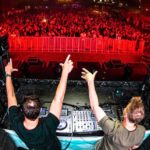 Everything You Wanted To Know About Trance (But Were Too Afraid To Ask) Two DJs perform on stage, raising their arms to a cheering crowd, illuminated by vibrant red lighting at a lively electronic music event.