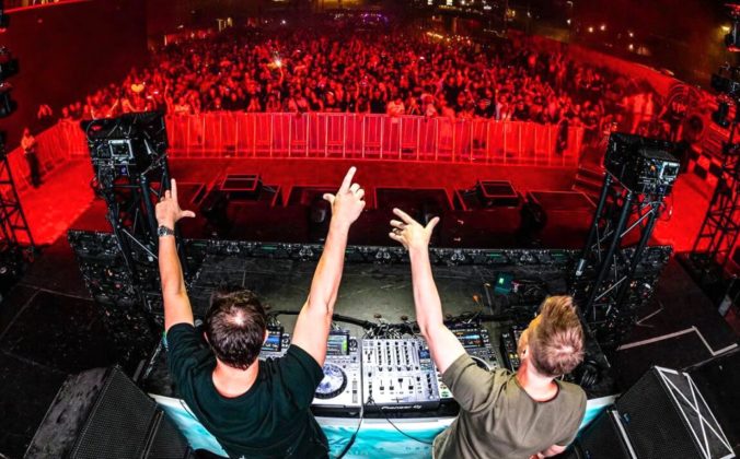 Two DJs perform on stage, raising their arms to a cheering crowd, illuminated by vibrant red lighting at a lively electronic music event.