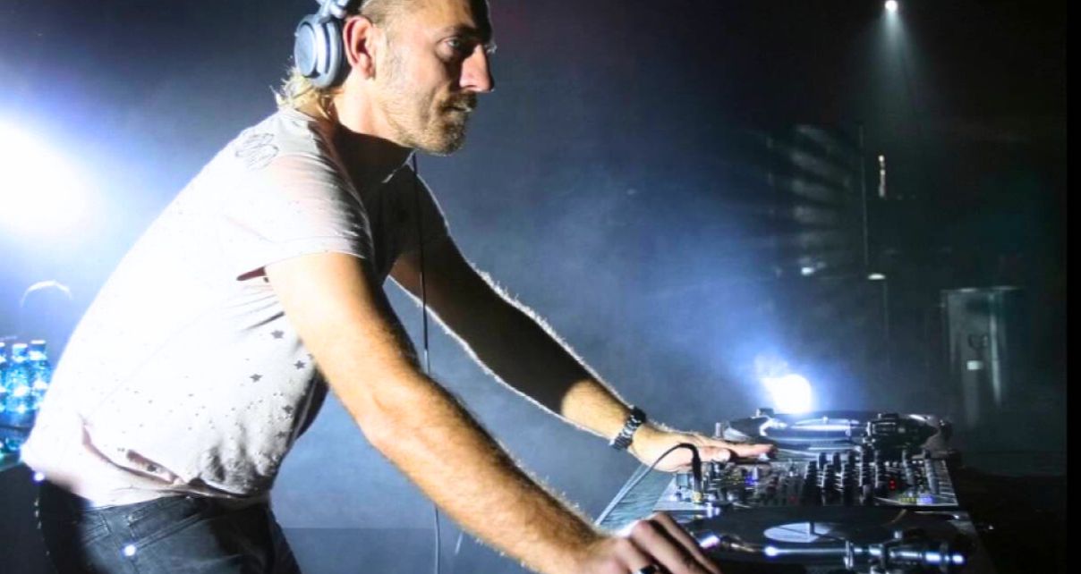 Sven Vath behind a pair of turntables in a darkened DJ booth, looking out at the crowd.
