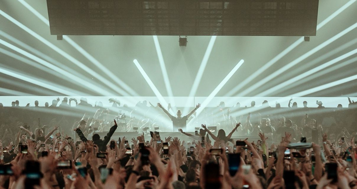 A vibrant festival scene with a Armin van Buuren performing on stage, surrounded by a cheering crowd beneath dramatic light beams.