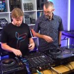 Phil Morse and Ferry Corsten stand behind a pro DJ set-up in a well-lit studio. They're both looking down towards the gear.