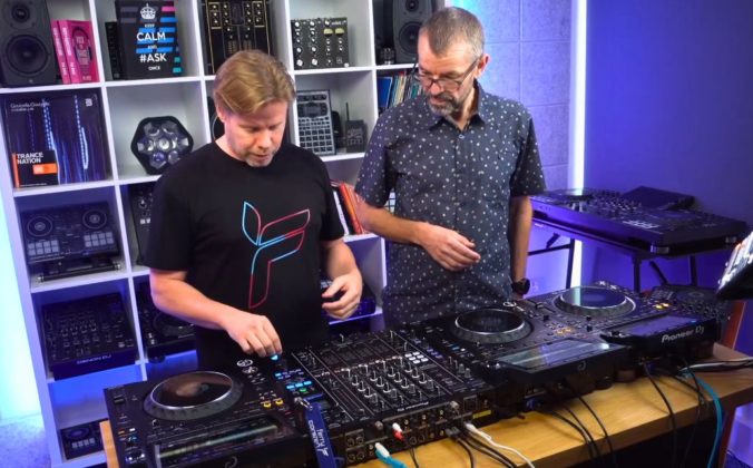 Phil Morse and Ferry Corsten stand behind a pro DJ set-up in a well-lit studio. They're both looking down towards the gear.
