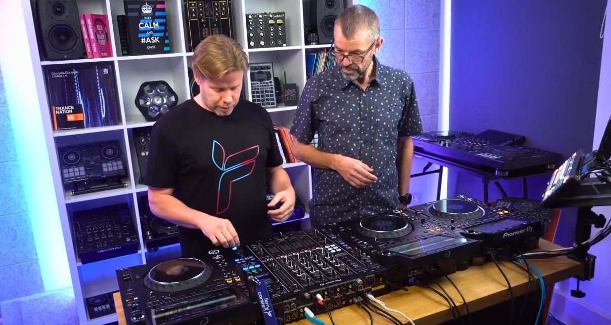 Phil Morse and Ferry Corsten stand behind a pro DJ set-up in a well-lit studio. They're both looking down towards the gear.