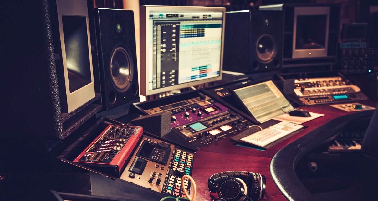 A professional music studio workspace featuring computer screens, speakers, audio equipment, and mixing tools. Headphones rest on the desk. The whole photo has a warm, vintage tone and feel.
