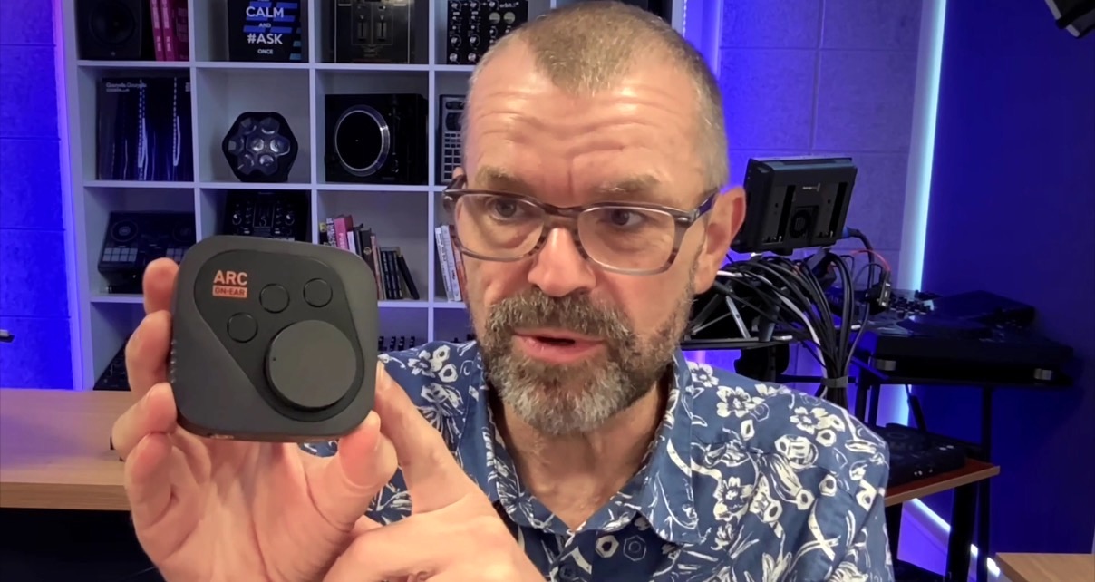 A bearded man with glasses sits in a well-lit studio. He's holding an Arc On-Ear device up to the camera and pointing at it.