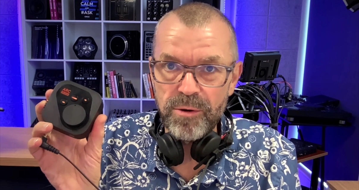 A bearded man with glasses and a blue and white patterned shirt holds up an Arc On-Ear box. He's in a well-lit music studio wearing black headphones.