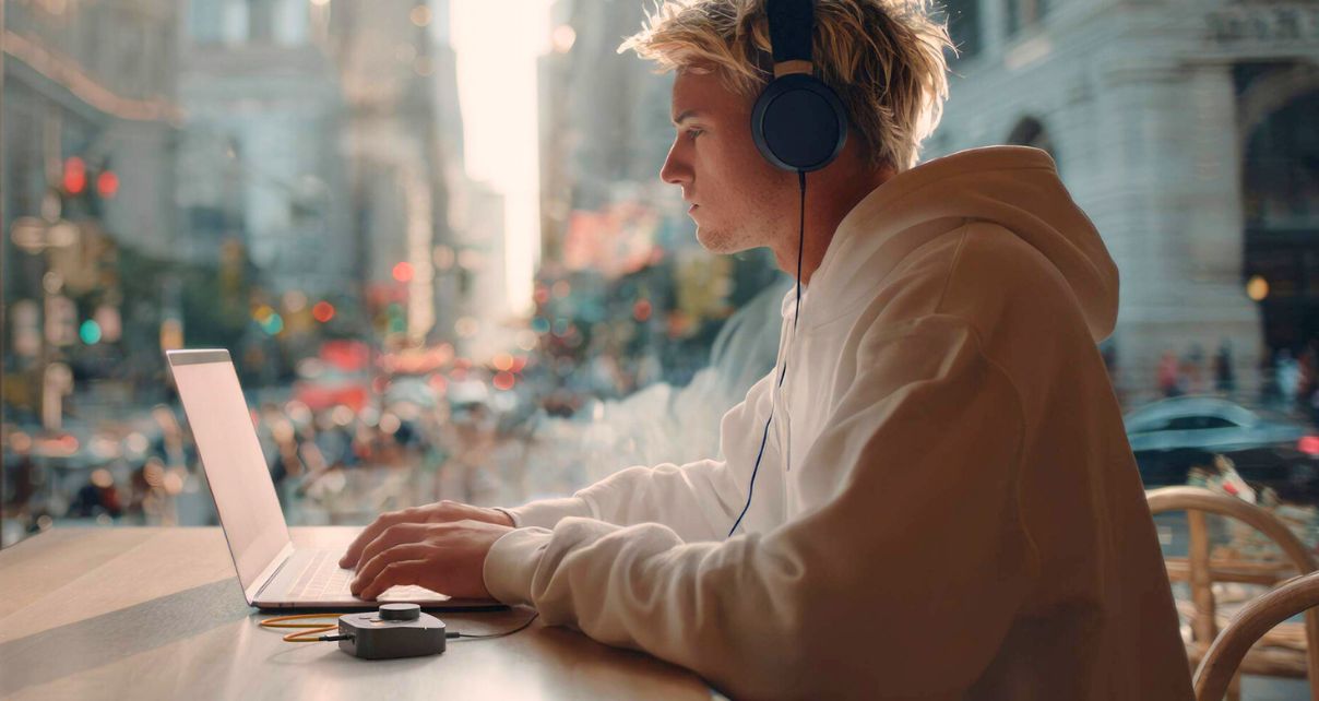 Side profile of a young blonde man working in headphones on a laptop. The room is warmly lit and he is sitting beside a window looking out into a city.
