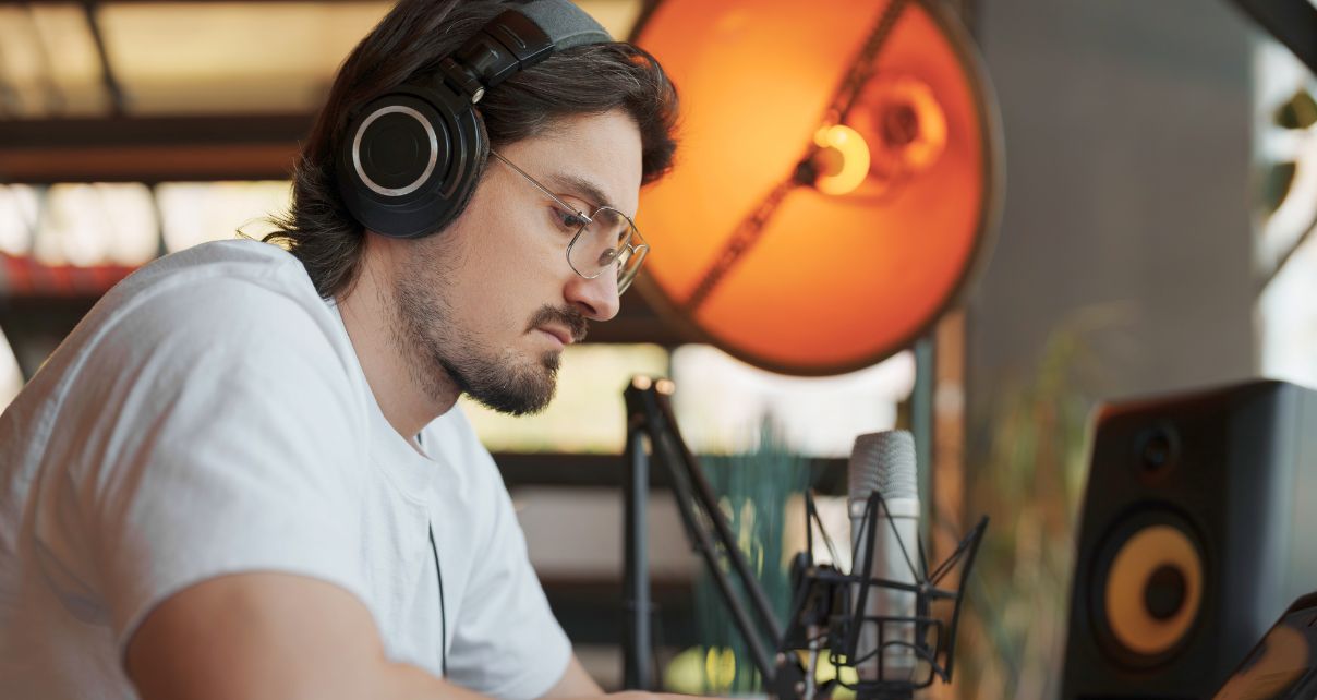 A man wearing black headphones and white t-shirt sits at a desk making music. The room is warmly lit and there is various music production gear in the background.