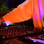 A DJ wearing a long sleeve white shirt and watch tweaks the mixer behind a DJ booth. It appears to be nighttime and the lighting is orange and purple.