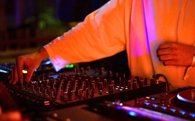 A DJ wearing a long sleeve white shirt and watch tweaks the mixer behind a DJ booth. It appears to be nighttime and the lighting is orange and purple.