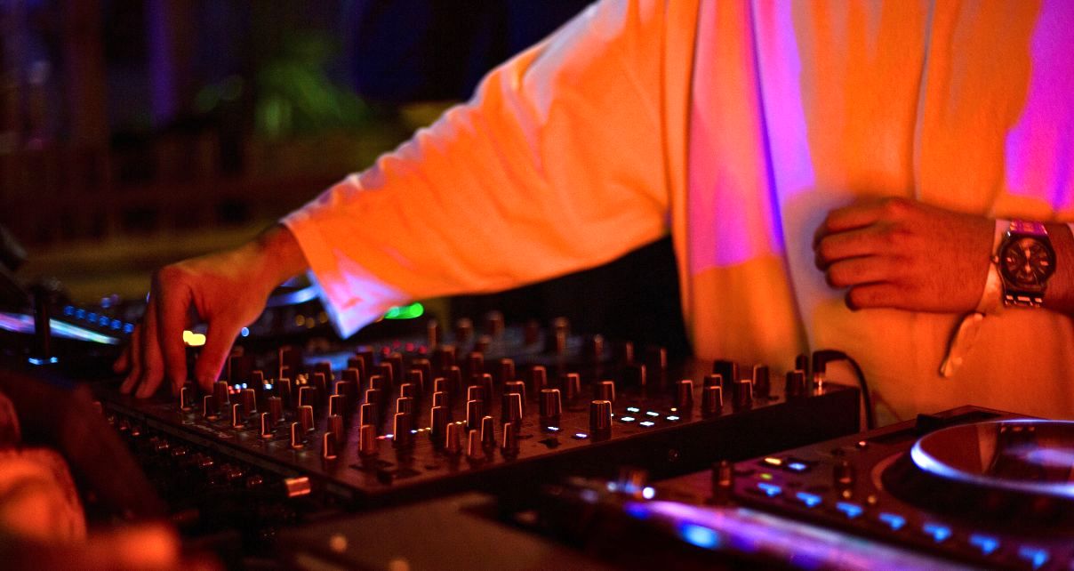A DJ wearing a long sleeve white shirt and watch tweaks the mixer behind a DJ booth. It appears to be nighttime and the lighting is orange and purple.