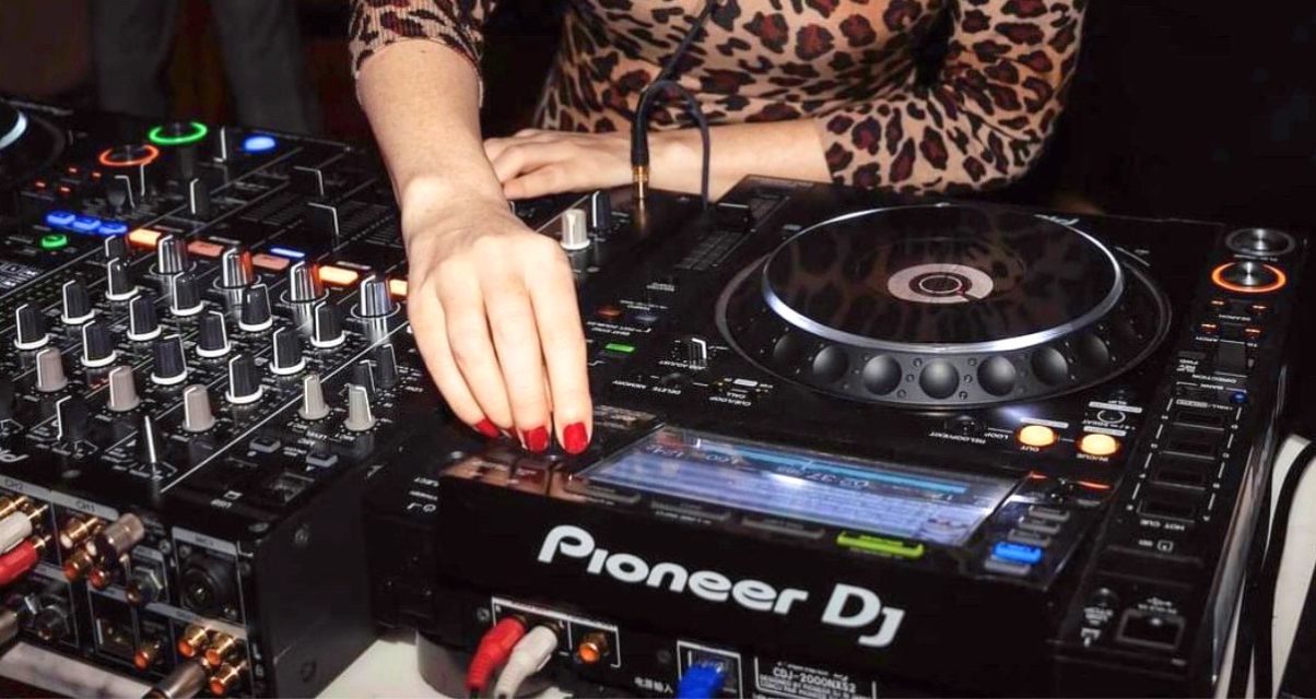 A DJ looks for a track on Pioneer DJ CDJs. They're wearing a leopard print shirt and have bright red nails.