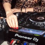 A DJ looks for a track on Pioneer DJ CDJs. They're wearing a leopard print shirt and have bright red nails.