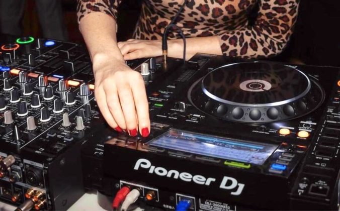 A DJ looks for a track on Pioneer DJ CDJs. They're wearing a leopard print shirt and have bright red nails.