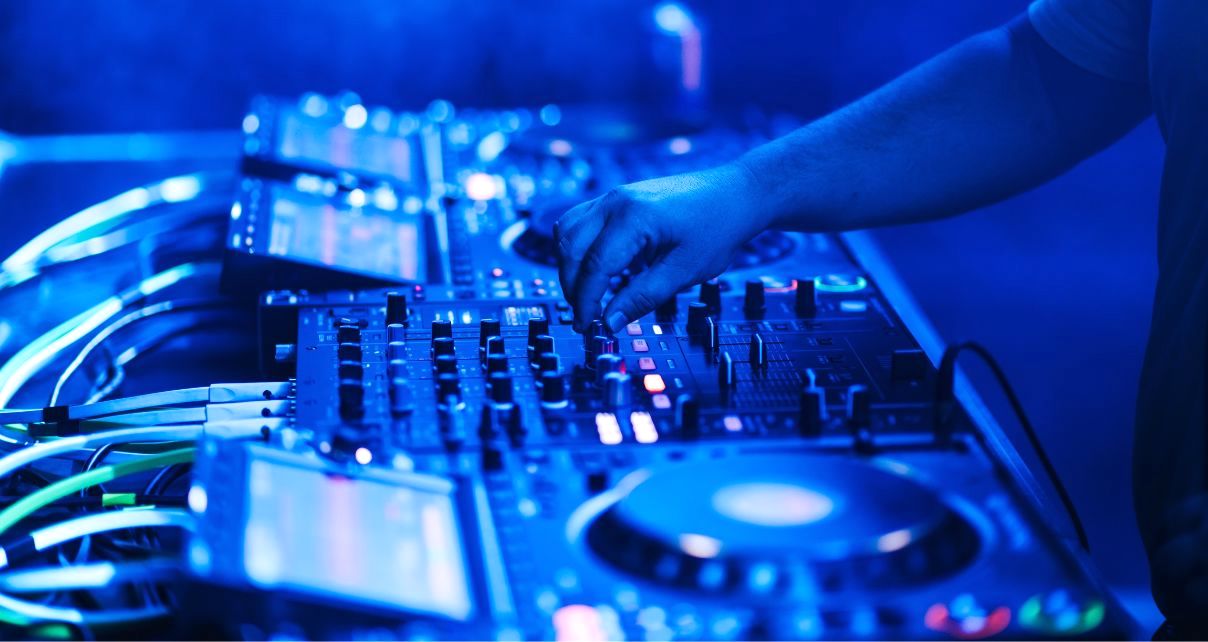 Close-up of a DJ mixing on three pro CDJs and a mixer. There are loads of cables plugged in around the back, and the whole scene has bluish lighting. 