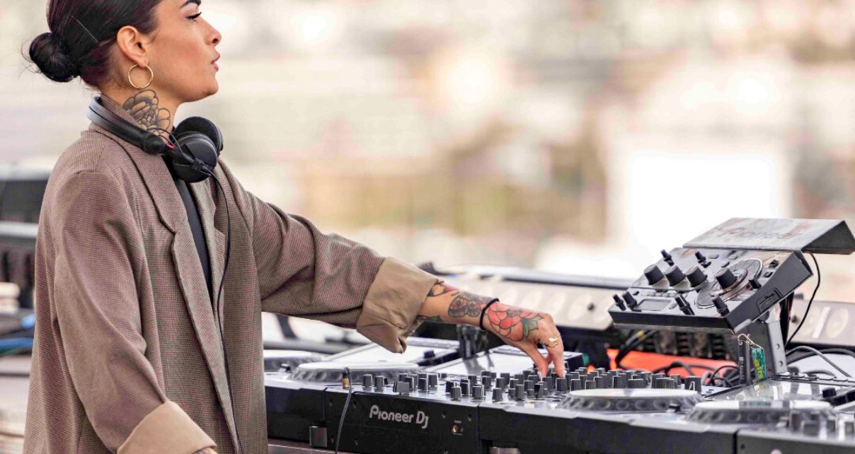 A feminine-presenting DJ with tattoos, hoop earrings, black headphones, and a large brown blazer looks at a festival crowd while DJing on CDJs. 